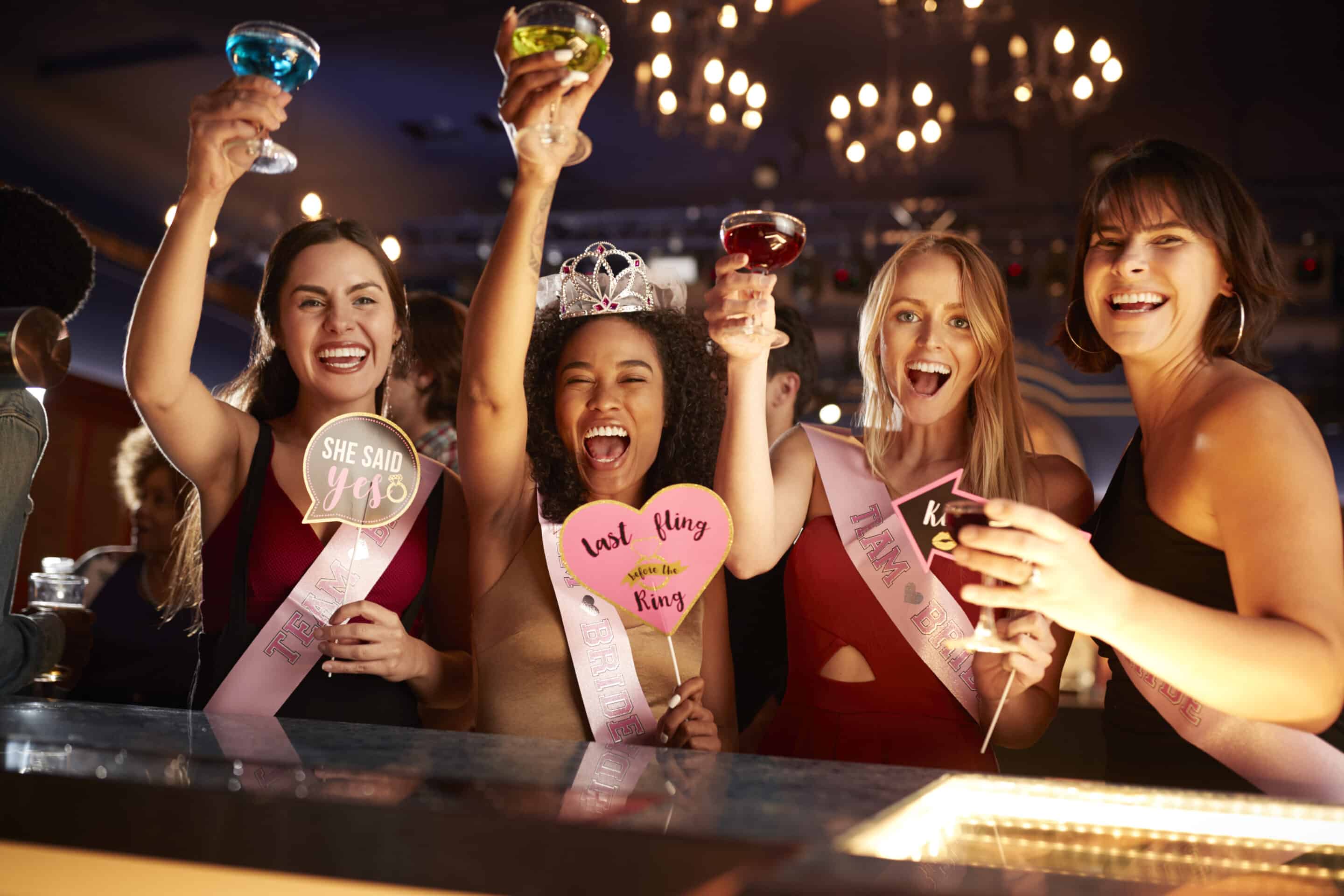 Four women at a bar celebrating a bachelorette party, smiling and raising drinks.
