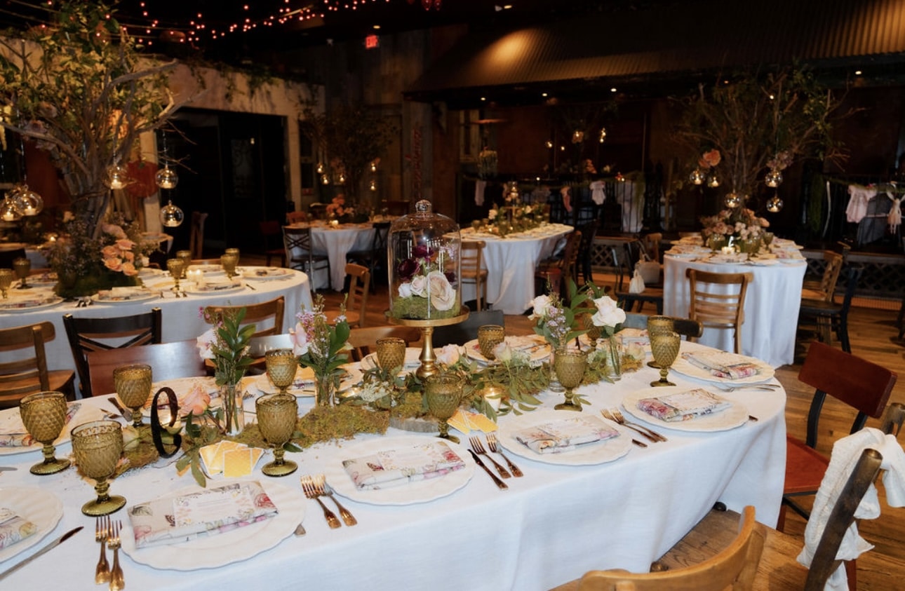 Long dining table set with white linens, amber goblets, floral centerpieces, and greenery runners in a warmly lit event space with string lights and round tables in the background.